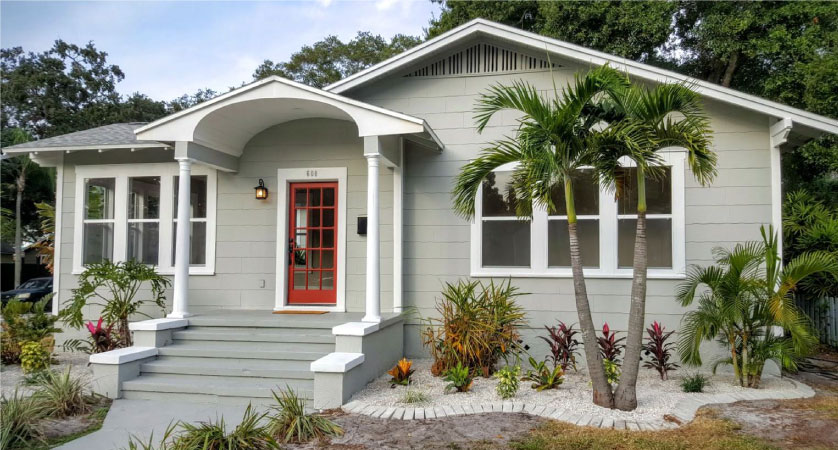 A one-story residential home in the Historic Kenwood neighborhood of St. Petersburg, Florida. The home is painted gray and white with a red front door. The front yard has a variety of tropical plants including palm trees. 
