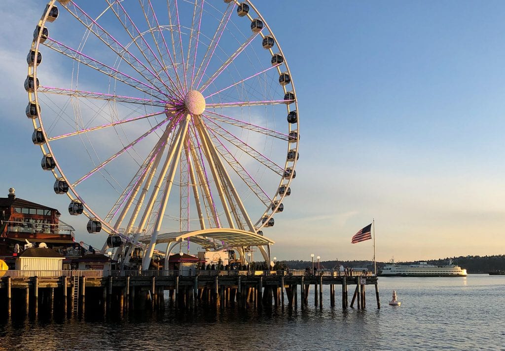 A close up of the Seattle Great Ferris Wheel and the peir it's on. There is a cruise ship in the background of the photo. 