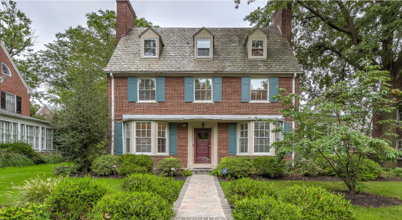 A stately brick home in Baltimore’s Guilford neighborhood is surrounded by mature trees, a lush green lawn, and decorative bushes, which are common in many nice Baltimore neighborhoods.