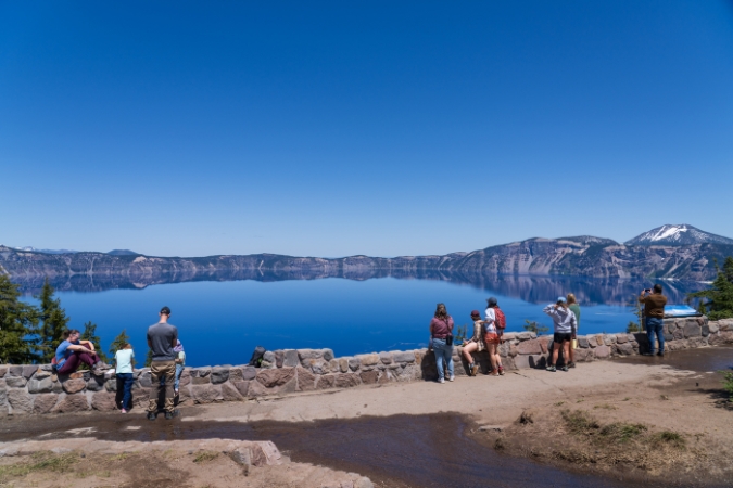Tourists admire Oregon’s Crater Lake with mountains and clear blue skies in the background.