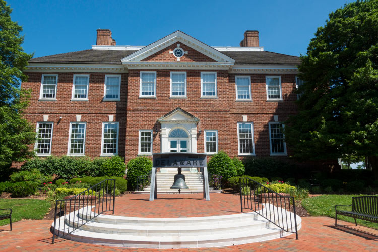 Exterior view of the Public Archives and Welcome Center at First State Heritage Park in Dover, Delaware. A large metal bell from the U.S.S. Delaware decorates the entrance, and a plaque above the bell reads “Delaware.”