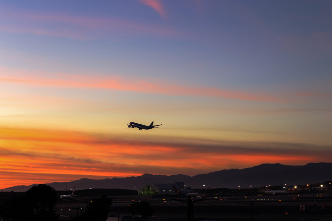 An airplane is departing from LAX during sunset with a vibrant orange and blue sky as a backdrop behind California mountains in the distance.