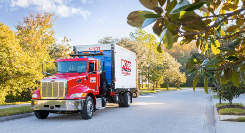 A PODS truck is transporting a PODS portable moving container through a residential neighborhood in Detroit on a sunny day.