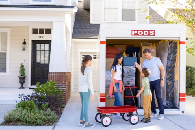 A family of four unloads boxes from a PODS portable moving container outside their new home, with a red wagon nearby.