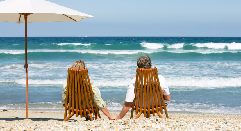 A retired couple lounges on two wooden chairs on a beach in Florida, one of the best states to retire. They’re holding hands and looking out at the surf of the blue Gulf waters.