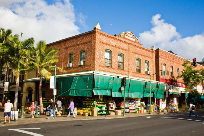 Street-level view of a brick building in Honolulu’s Chinatown, featuring green sun shades and stacks of fresh produce.