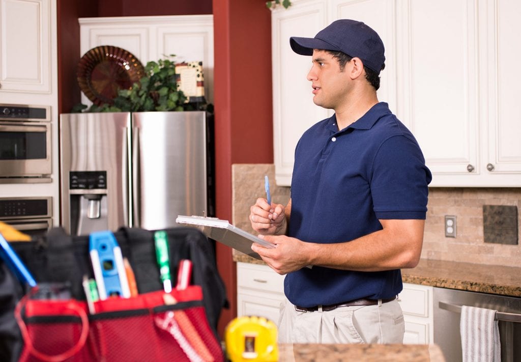 a contracotr in a navy plo and hat makes a checklist of things to inspect. his bag with a tape measurer sits on a granite counter. 
