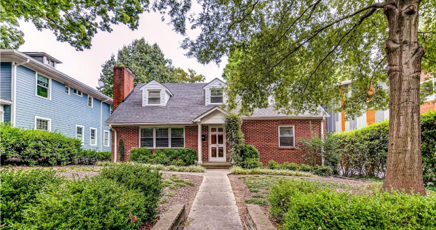 A cottage-like smaller brick home with three dormers and a covered porch sits between two larger, multi-story houses in Forest Park.