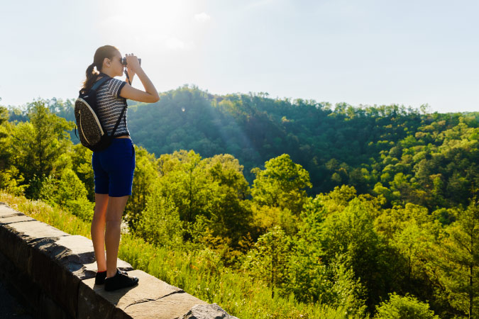 A young woman is standing on a stone wall, using binoculars to look out over Cherokee National Forest in Tennessee on a sunny day.