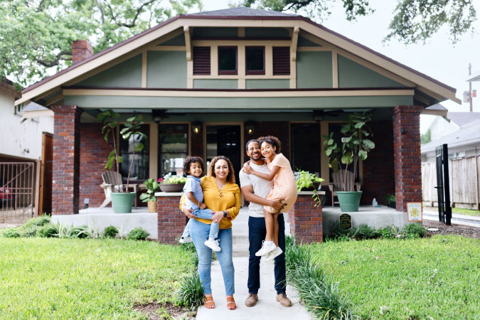A happy family of four poses together for a photo outside their new green and brick bungalow-style home in Texas.