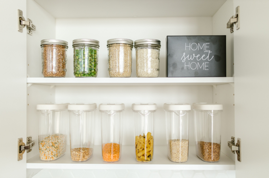 A close up of jars and storage containers in a neatly organized kitchen cabinet. There is a tiny sign to the right that reads "home sweet home".