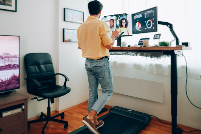 A man is walking on a floor treadmill at his standing desk, which features two large monitors — all items that were on his home office wish list.