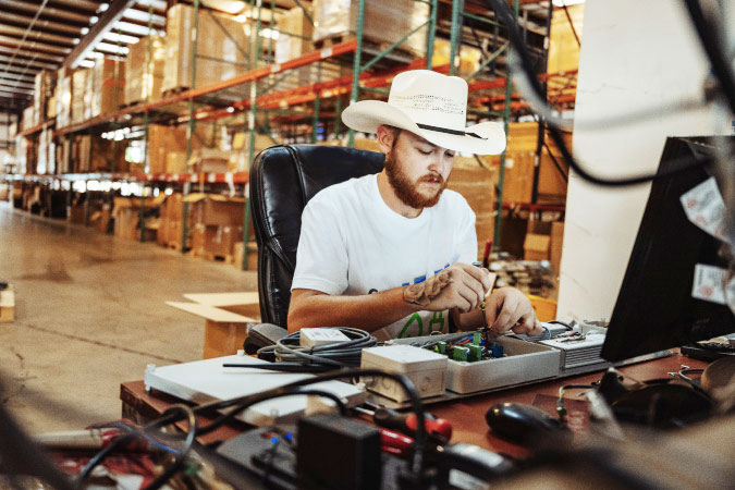 A young man in a cowboy hat is working at a computer station in a warehouse in Texas.
