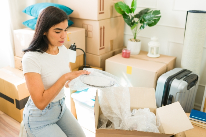 A woman is demonstrating how to pack dishes for moving, wrapping a plate with bubble cushioning roll before placing it inside of a moving box.