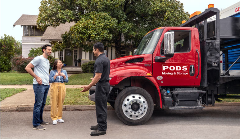 A couple and a PODS truck driver stand in the street in front of a PODS moving truck. 