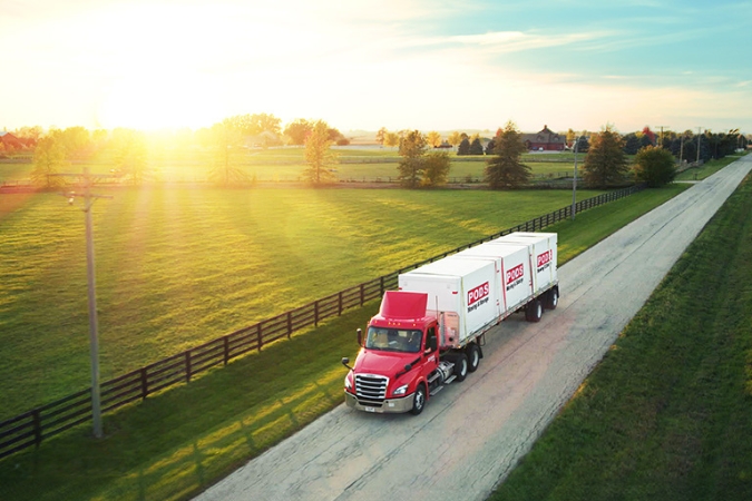 As part of a residential move down South, a PODS truck is transporting three portable moving containers along a rural road lined with green pastures.