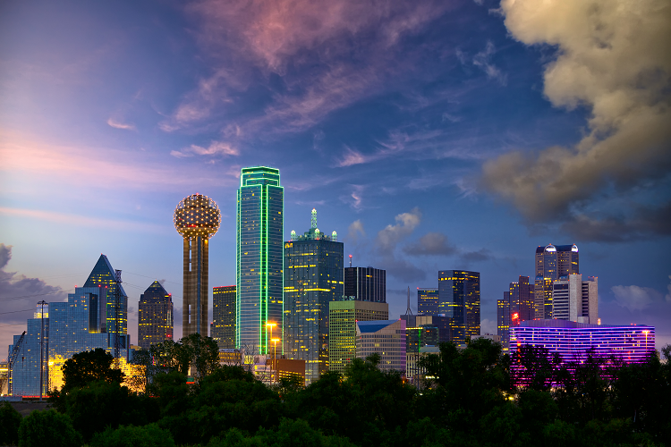The Dallas, Texas, skyline at dusk. There are high-rise buildings illuminated with multi-colored lights that complement the night sky.
