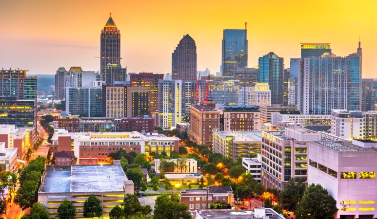Aerial view of Atlanta, Georgia, at sunset. City lights have begun to come on, and the tall downtown buildings create a distinct skyline against a yellow sky.