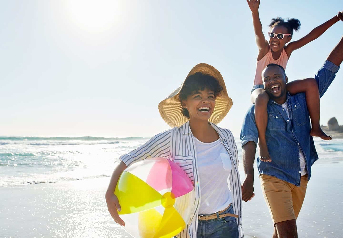 A mom, dad, and daughter smiling on the beach. Daughter sits on dad's shoulders while Mom carries a beach ball