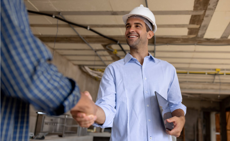 A man in a white hard hat and blue button up shirt holds a clipboard in one hand and shake the hand of a man in a blue checked shirt with the other. 