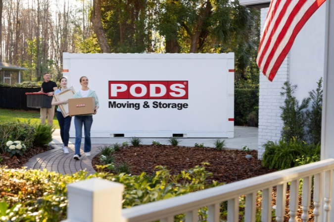 A military family moving into their new home is carrying moving boxes from a PODS portable moving container toward the front door.