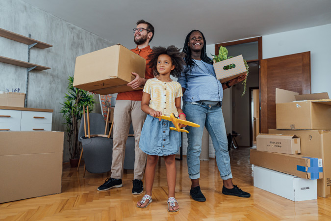 A happy family of three, with the mother expecting, is standing in their new home surrounded by moving boxes and miscellaneous household items.