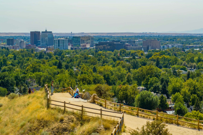 A vibrant, wide-angle view of Boise, Idaho, with a paved walking trail filled with dense green trees in the foreground. In the background, the downtown skyline is visible.