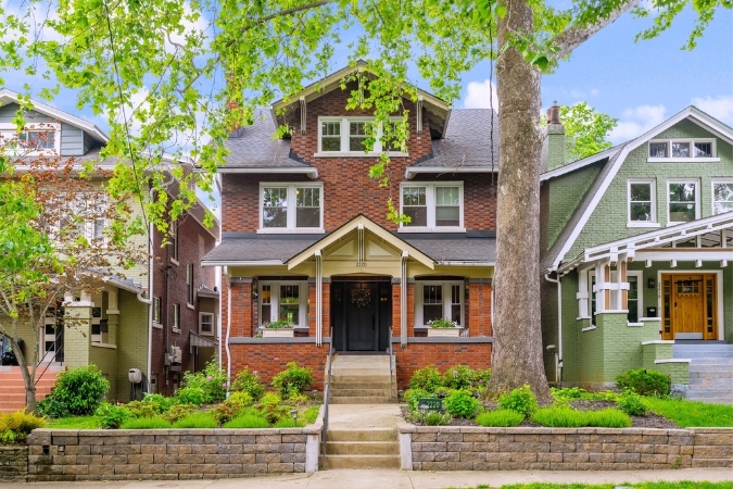 A red brick, three-story American Craftsman–style home stands between neighboring houses, with a central staircase dividing a shrub-dotted front lawn.