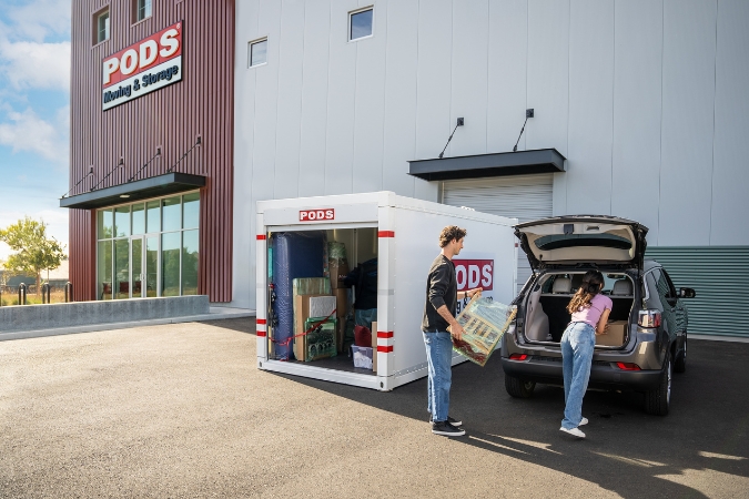 A young couple in the parking lot of a PODS Storage Center, moving belongings to the trunk of their car from a loaded PODS portable storage container — one of the most flexible moving and storage options.