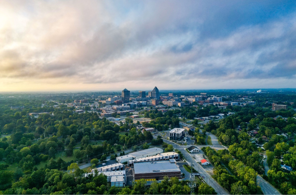 An aerial view of downtown Greensboro, highlighting what it's like living in Greensboro, NC, with lush green spaces and tall buildings under a partly cloudy morning sky.