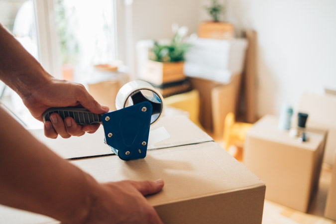 Close-up of a pair of hands using packing tape to seal a moving box, with stacks of moving boxes and other household items in the background.
