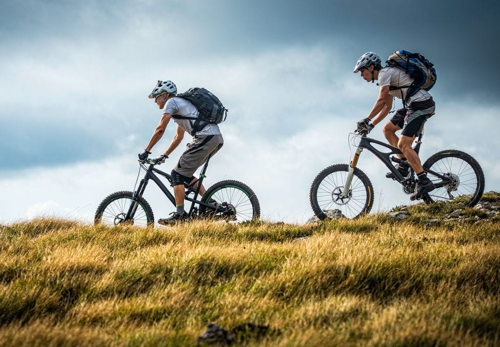 Two middle-aged men have backpacks on their backs and ride bikes in the mountains.
