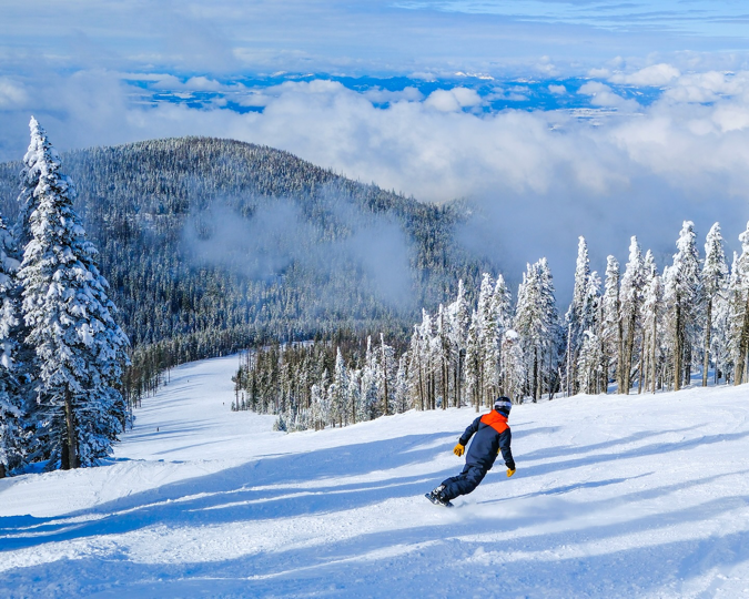A snowboarder living in Spokane, WA, descends a mountainside dotted with snow-covered trees on a bright, partly cloudy winter day.