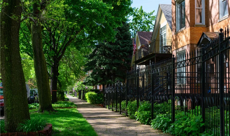 View down a tree-lined sidewalk in Chicago’s Logan Square neighborhood, featuring brick townhomes behind decorative iron gates.