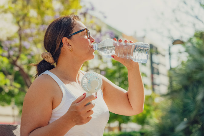 A middle-aged woman is holding a small electric fan and drinking a bottle of water on a hot, sunny day while living in Dallas, Texas.