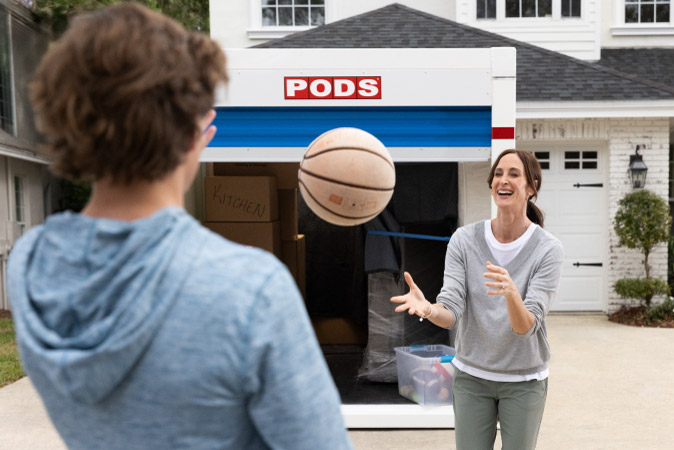 A mother and son toss a basketball back and forth in front of a loaded PODS portable moving container in the driveway of their new home.