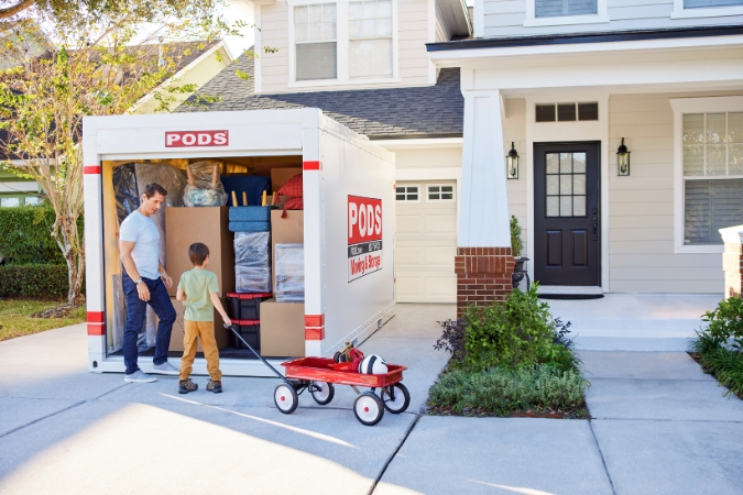 A father and son moving to Greensboro, NC, stand outside of their loaded PODS moving container as the son pulls a red wagon filled with some household items.