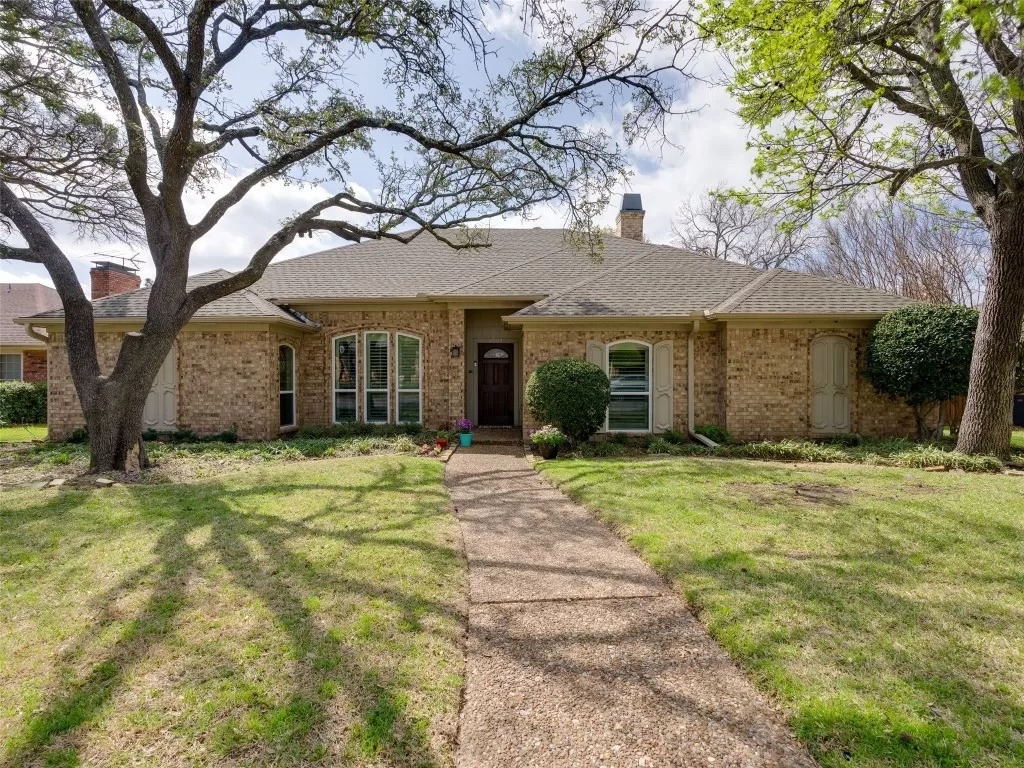 A brick one-story home in the neighborhood of Preston Highlands in Dallas, Texas. It is framed by two tall oak trees in a green lawn. There is a sidewalk with the house number painted on the curb. 
