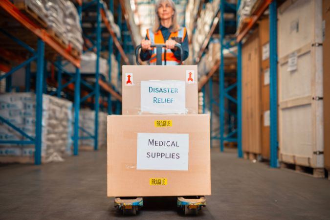 A facilities worker is moving boxes labeled “DISASTER RELIEF” and “MEDICAL SUPPLIES” through a warehouse to be stored for emergency response purposes.