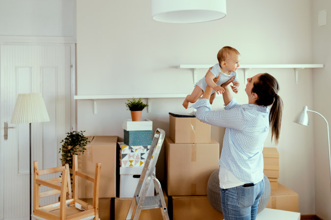 While preparing for moving with a baby, a mom holds her infant playfully above her head in a room filled with moving boxes, a ladder, and a few pieces of furniture.