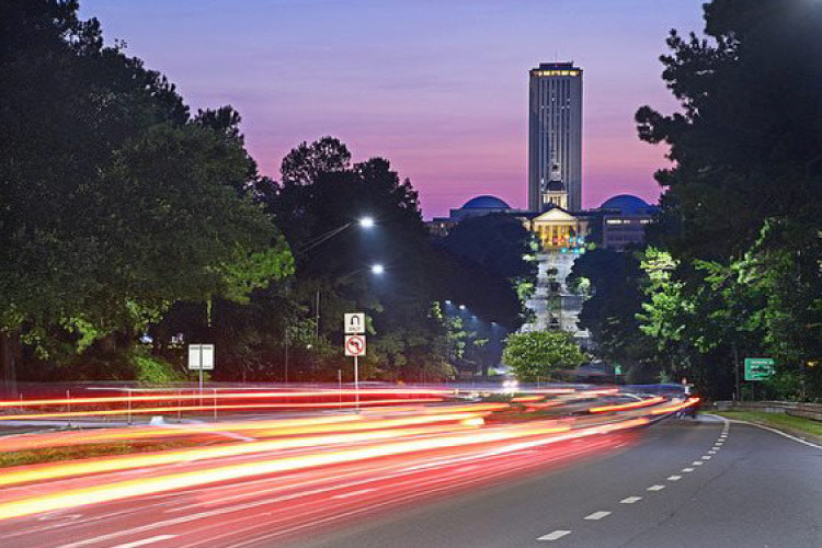 Traffic zips along Tallahassee’s Highway US-27 at dusk with the city’s capitol building in the background. The cars are all a blur of red and yellow lights.