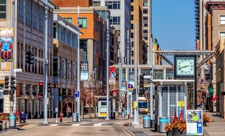 Street-level view of an intersection in downtown Minneapolis, featuring commercial buildings and a bus stop with a large clock face on display. 