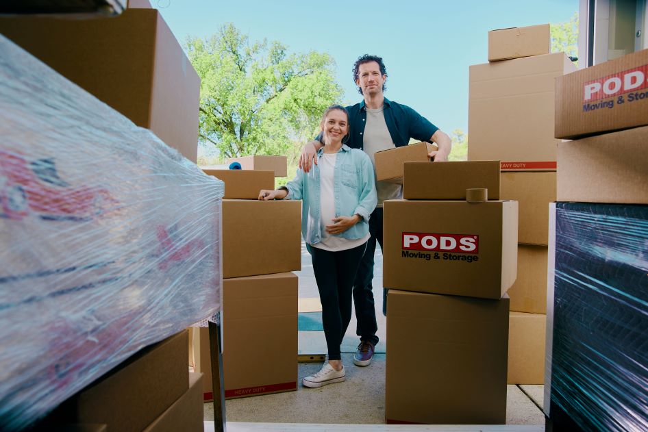 A young, pregnant couple navigates moving day chaos, standing in front of boxes as they prepare for their move.