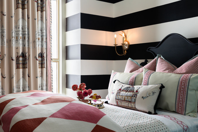 A newly redesigned bedroom showcasing high contrast home remodeling ideas, including black-and-white striped walls, dramatic drapes, gold wall sconces, and a colorful bedset.