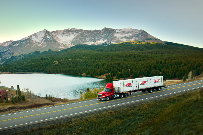 A PODS truck transporting three portable moving containers along an interstate road, past a lake, forest, and distant mountains.