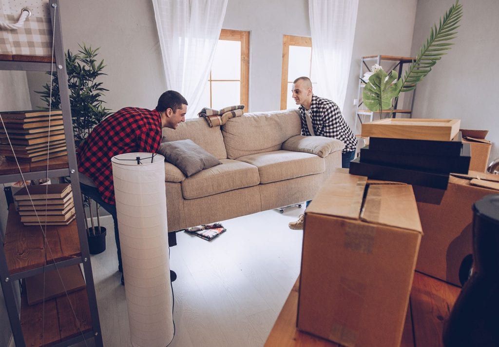 Two men, both wearing flannel are lifting a tan couch. Ther are moving cardboard boxes everywhere.