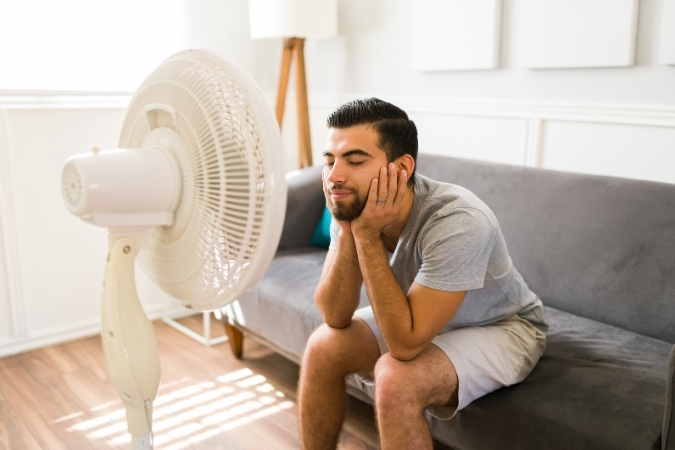 A man sits on a couch in front of a fan to cool off while following tips for moving in the summer.