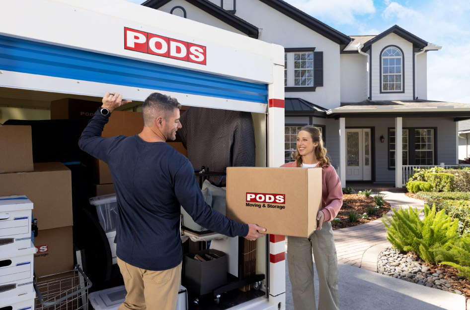 A woman is handing a PODS branded moving box to her husband to load into the nearly full PODS portable moving container in their driveway.