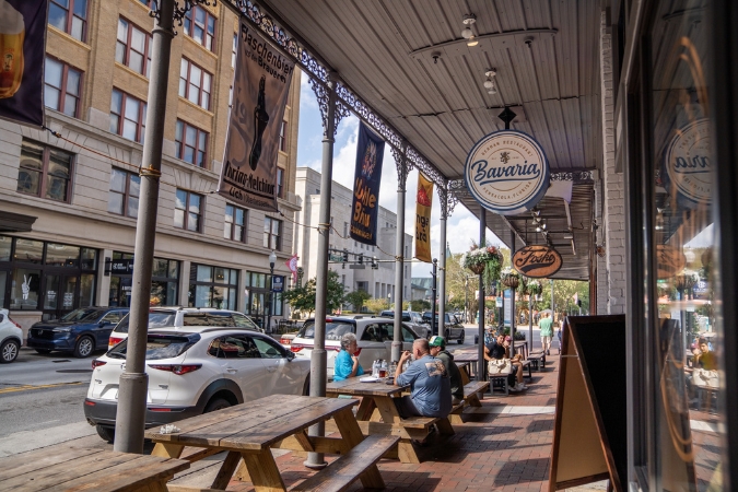 A group of people living in Pensacola are dining al fresco at a Downtown restaurant on a bustling street lined with shops and historic buildings.