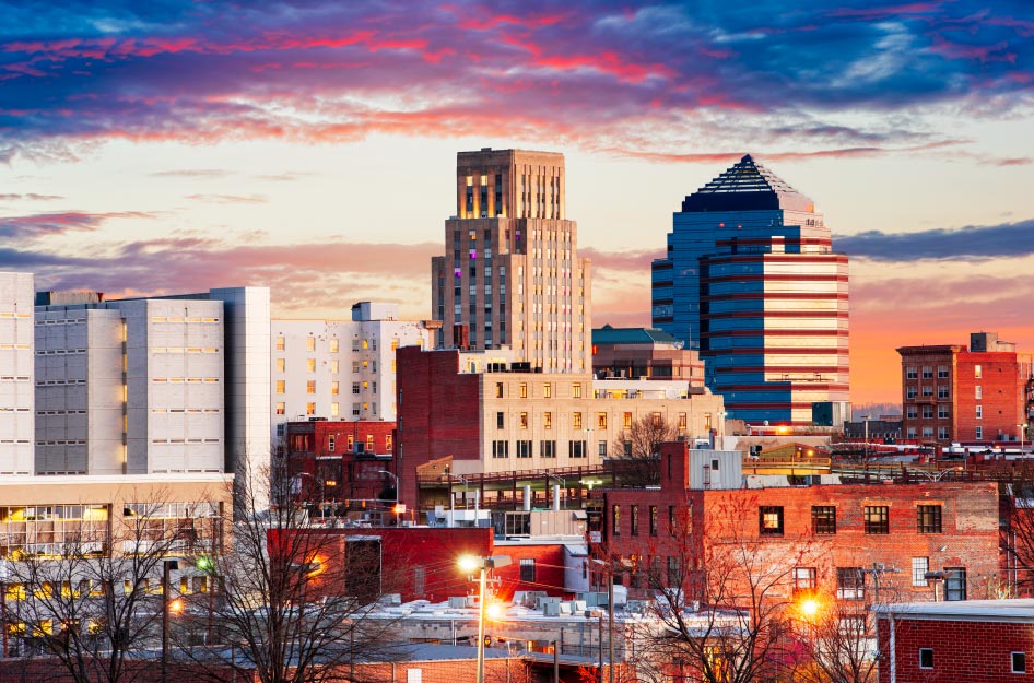 Sunset view of the Durham, NC, skyline with illuminated street lights below and vibrant pink and purple clouds above.